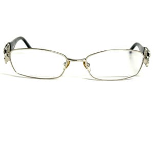 May include: A pair of silver-toned metal eyeglasses with rectangular lenses. The frames feature thin, metallic rims and dark-colored temple arms. The bridge connects the two lenses, and the hinges display decorative metallic detailing on each side.