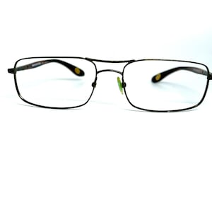 May include: Rectangular eyeglasses with a thin, dark metallic frame and a double bridge design. The temples feature dark tips with small, oval-shaped yellow accents. The nose pads are translucent and have a slight greenish tint.