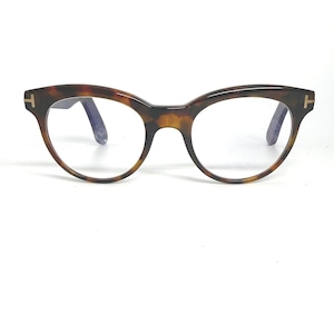 Może przedstawiać: Okulary w kształcie kocich oczu z wzorem w kolorze żółwia. Oprawki mają brązowy i beżowy wzór, ze złotym logo 'T' na każdej zauszniku. Soczewki są przezroczyste.