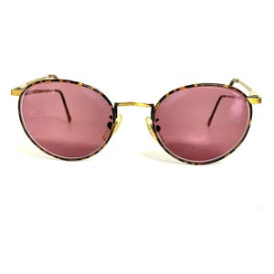 May include: Round eyeglasses featuring pink-tinted lenses and a tortoiseshell pattern frame. The glasses have thin gold-tone metal temples and a matching bridge, with adjustable nose pads visible behind the lenses, creating a classic eyewear look.