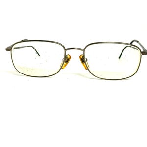 May include: A pair of rectangular eyeglasses with a thin, dark metallic frame. The glasses feature clear lenses and amber-colored nose pads. The bridge connects the two lenses, and the thin temples extend outward from the outer edges of the frame.