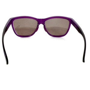 May include: Purple sunglasses with a rectangular frame and dark lenses. The arms are black and curve away from the frame. The sunglasses are set against a white background.