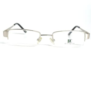 Pode incluir: Óculos com armação prateada de formato retangular e lentes transparentes. As hastes são pretas e a marca "di caprio" está impressa na lente direita. Os óculos estão sobre um fundo branco.
