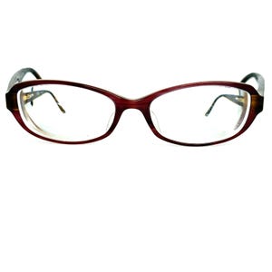 Pode incluir: Um par de óculos com armação bordô e lentes transparentes. A armação tem formato arredondado com efeito degradê, passando do bordô para uma cor mais clara nas hastes. As hastes são marrons escuras.