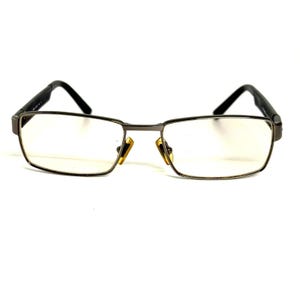 Pode incluir: Óculos retangulares com armação de metal cinza-escuro e lentes transparentes. O design inclui hastes finas e retas, além de plaquetas nasais douradas, com uma ponte que conecta os dois aros das lentes.