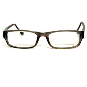 Pode incluir: Óculos retangulares com uma armação cinzento-escura translúcida com um subtil tom castanho. Os óculos têm lentes transparentes e hastes finas e curvas que se estendem a partir dos cantos exteriores da armação, num design simples e funcional.