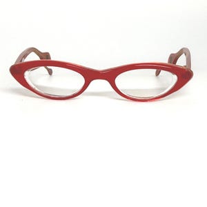 May include: Red cat-eye style eyeglasses with clear lenses. The frames are a vibrant red, with a slightly translucent appearance. The temples are a contrasting brown color. The glasses are set against a white background.