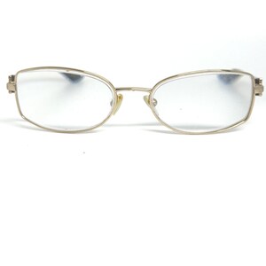 May include: Gold-toned eyeglasses with a rectangular lens shape. The frames have a thin, delicate design with a bridge and nose pads. The lenses are clear, and the glasses appear to be in good condition, suitable for vision correction.