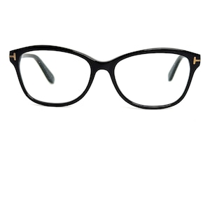 以下が含まれることがあります： 黒いキャットアイフレームの眼鏡。各テンプルには金色の「T」ロゴがあります。レンズは透明で、フレームは光沢のある黒色です。白い背景に配置されています。