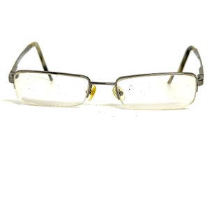 May include: Rectangular eyeglasses featuring a thin, dark metallic frame. The glasses have clear lenses and small, yellow-tinted nose pads attached to the bridge. The temples extend outward from the sides, completing the structure of these spectacles.