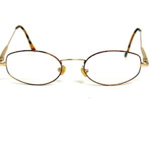 May include: Oval-shaped spectacles with a gold-coloured metal frame. The arms have a tortoiseshell pattern. The lenses are clear. The nose pads are a light yellow colour. The glasses are set against a white background.