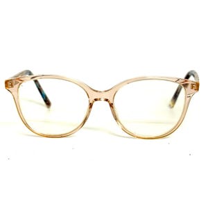 May include: Clear, translucent peach-toned eyeglasses with a rounded frame shape. The temples feature a subtle, multi-colored speckled pattern in shades of blue, brown, and white, visible through the transparent material of the frame arms.