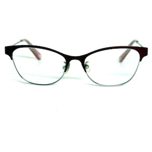 May include: Cat-eye style eyeglasses featuring a dark maroon upper frame and a thin silver-toned lower rim. The glasses include clear lenses, adjustable nose pads, and slender temples with pinkish-toned tips, presenting a classic semi-rimless design.