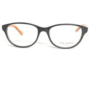 May include: Black eyeglasses with a cat-eye shape and orange accents on the temples. The glasses have clear lenses and the brand name "TED BAKER LONDON" is printed on the right lens.