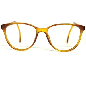 May include: A pair of amber-colored eyeglasses with a translucent, rounded frame. The glasses feature a smooth, polished finish and thin, curved temples that extend outward. The lenses are clear and unobstructed, showcasing the simple, classic design.