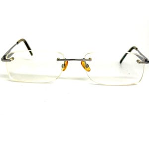 May include: Rimless eyeglasses featuring rectangular clear lenses and a thin, dark metallic bridge. The frame includes slender dark metal temples and amber-colored nose pads, providing a lightweight and streamlined appearance for these optical glasses.