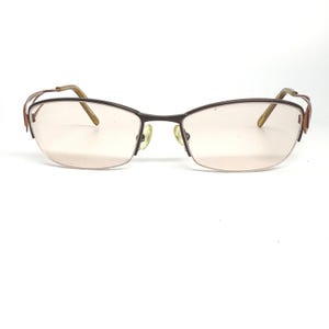 May include: A pair of eyeglasses with a dark brown metal frame and light pink lenses. The glasses have a semi-rimless design with a curved top and a straight bottom. The temples are a lighter brown color.