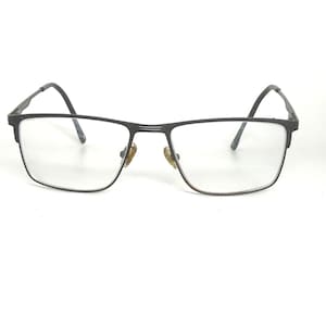 Op de afbeelding: Rechthoekige bril met een donkergrijs metalen montuur. De glazen zijn helder en de pootjes buigen om de oren. De bril heeft een klassiek, minimalistisch ontwerp.