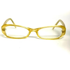 May include: A pair of spectacles with a translucent, light yellow, horn-rimmed frame. The glasses feature an elongated, slightly oval lens shape with thin, curved temples extending from the sides. The frame has a smooth, polished finish.