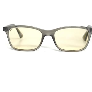 May include: Gray-framed eyeglasses with yellow-tinted lenses. The rectangular frames have a slightly rounded shape and a matte finish. The lenses are clear with a yellow hue, suggesting they may be designed to filter blue light.