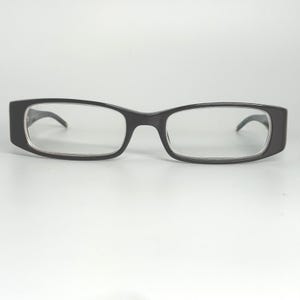 May include: Black rectangular eyeglasses with clear lenses. The frames are thick and dark, with a slight curve at the temples. These glasses are designed for vision correction and are suitable for daily wear.