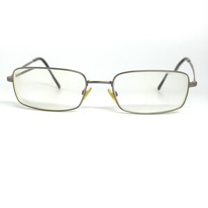 May include: Rectangular eyeglasses with a thin, silver-gray metal frame. The lenses are clear, and the temples are dark gray. The glasses have a classic, minimalist design, suitable for everyday wear. The frame is approximately 5.5 inches wide.