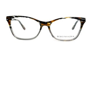 May include: BCBGMAXAZRIA eyeglasses with a cat-eye frame. The frames feature a gradient design, transitioning from a light gray at the bottom to a tortoiseshell pattern at the top. The lenses are clear, and the temples are a dark gray.