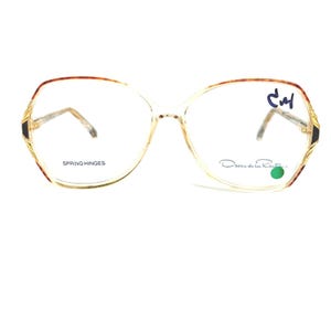 May include: Clear-framed eyeglasses with a geometric shape. The frames have gold accents and brown detailing on the upper rims. The text "SPRING HINGES" is printed on the left lens, and the brand name "Oscar de la Renta" is printed on the right lens.