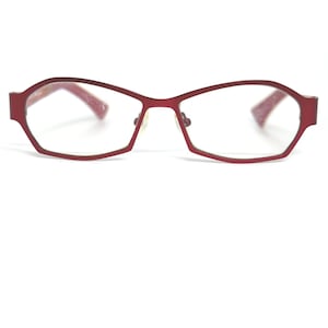 May include: Red eyeglasses with a geometric frame design. The glasses have a rectangular shape with rounded edges and clear lenses. The temples are also red and have a textured appearance. The nose pads are a light color.