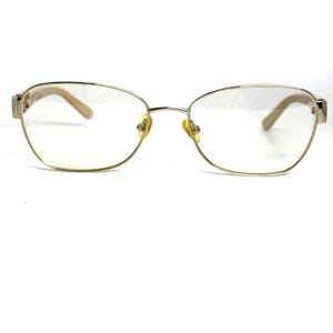 May include: Gold-tone eyeglasses with a delicate wire frame and oval lenses. The temples and nose pads are a light beige color. The glasses have a classic, elegant design.