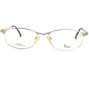 May include: Gold-coloured eyeglasses with a rectangular frame and black temples. The lenses are clear, and the frame has the text "MONEL ITALY DESIGN" and "De Moda". The glasses have a decorative design on the temples.