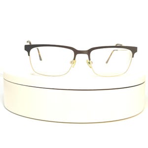 Pode incluir: Óculos com armação superior castanho escuro e inferior dourada. As lentes são transparentes e os óculos têm um formato retangular clássico. Os óculos estão sobre um fundo branco.