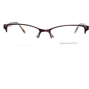 May include: A pair of eyeglasses with a dark purple metal frame. The glasses have a curved browline and a thin, delicate design. The brand name, BCBGMAXAZRIA, is printed on the right lens.