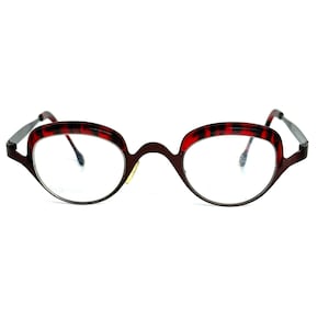 Pode incluir: Óculos ovais com armação castanha escura e detalhes estampados em vermelho e preto nas bordas superiores. As lentes são transparentes e as hastes são prateadas. Os óculos têm uma estética vintage.