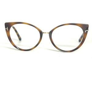 Może przedstawiać: Okulary w kształcie kocich oczu z wzorem w żółwiej skorupie. Oprawki mają brązowo-beżowy wzór ze złotym mostkiem i zawiasami. Soczewki są przezroczyste, a okulary mają klasyczny, stylowy wygląd.