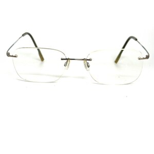 May include: Rimless eyeglasses featuring thin, metallic silver-toned temples and a matching bridge. The clear, rectangular lenses are secured by small metal hardware at the bridge and outer edges, complemented by dark-colored temple tips.