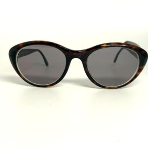 May include: Oval-shaped sunglasses featuring a dark brown and black tortoiseshell pattern frame. The lenses are a solid dark grey tint, providing a neutral appearance. The frame has a smooth, rounded bridge and thin, dark temples extending outward.