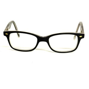 Pode incluir: Um par de óculos com uma armação frontal em preto brilhante e um aro interior transparente. As hastes também são transparentes. Pequenos detalhes decorativos de cor dourada em forma oval são visíveis nos cantos externos da armação.