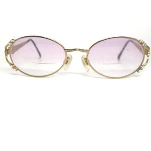 May include: Gold-tone oval eyeglasses with gradient pink lenses. The frames feature decorative accents with small clear stones on the temples. The nose pads are a light beige color.
