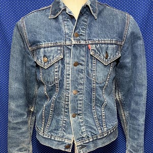 May include: A vintage blue denim jacket with a classic design. The jacket features two chest pockets, button closures, and a red tag. The jacket is displayed on a mannequin against a blue background.