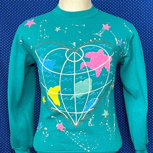 May include: Teal sweatshirt with a white heart-shaped globe design. The globe is surrounded by white waves and pink, yellow, and blue continents. The sweatshirt is decorated with white paint splatters and pink, yellow, and blue stars.