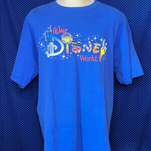 May include: A blue t-shirt featuring the "Walt Disney World" logo in a colorful design. The graphic includes a castle, fireworks, and the iconic Mickey Mouse ears. The shirt is displayed on a mannequin.