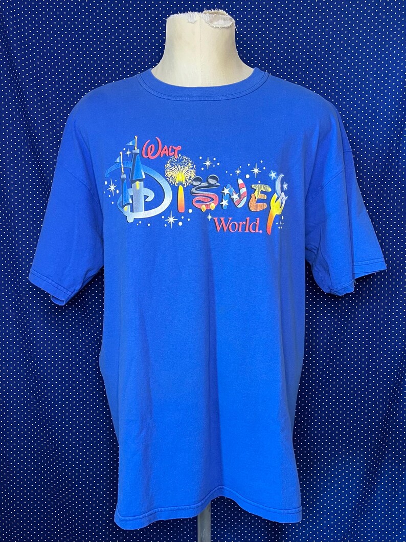 May include: A blue t-shirt featuring the "Walt Disney World" logo in a colorful design. The graphic includes a castle, fireworks, and the iconic Mickey Mouse ears. The shirt is displayed on a mannequin.