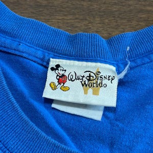 May include: Close-up of a blue t-shirt with a Walt Disney World tag. The tag features Mickey Mouse and the words "Walt Disney World" in a stylized font. The shirt's fabric is visible, showing its texture.