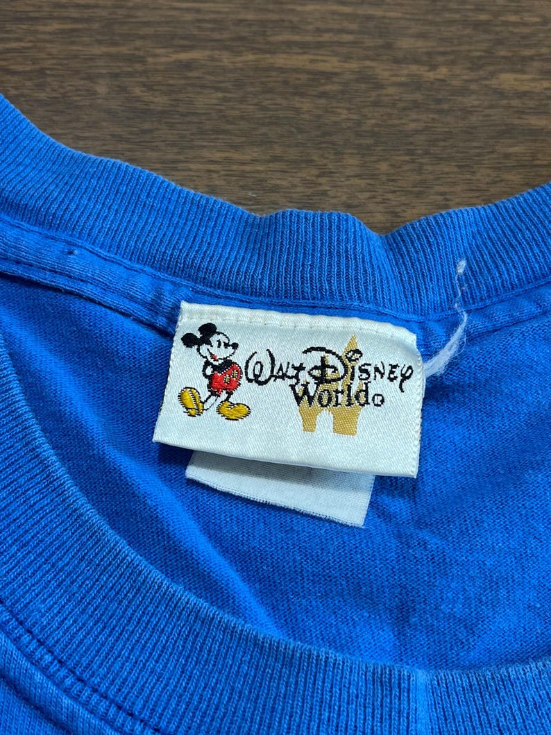 May include: Close-up of a blue t-shirt with a Walt Disney World tag. The tag features Mickey Mouse and the words "Walt Disney World" in a stylized font. The shirt's fabric is visible, showing its texture.