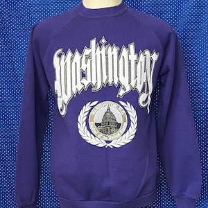 May include: Purple sweatshirt with a white graphic of the word "Washington" in a gothic font. The graphic also includes a white and gold emblem with the words "Washington D.C. Capital of the United States" and a depiction of the Capitol Building.