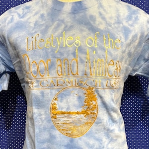Puede incluir: Una camiseta de color azul claro tie-dye con letras doradas que dicen "Lifestyles of the Poor and Aimless" y un círculo dorado con un lago y árboles en su interior. El texto "Of Garmisch USA" está debajo del círculo.
