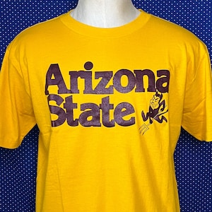 Könnte beinhalten: Gelbes T-Shirt mit dem Schriftzug "Arizona State" in braunen Blockbuchstaben. Auf der rechten Seite des Textes befindet sich ein brauner Cartoon-Teufel mit einer Mistgabel. Das Shirt wird auf einer Schaufensterpuppe präsentiert.