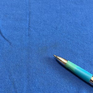May include: Close-up of a blue t-shirt with the word "World" in red and orange. A pen with a teal and silver barrel is in the foreground, pointing towards the center of the shirt.