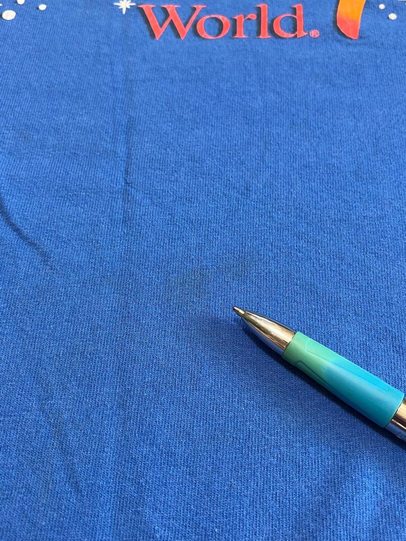 May include: Close-up of a blue t-shirt with the word "World" in red and orange. A pen with a teal and silver barrel is in the foreground, pointing towards the center of the shirt.